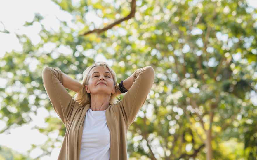 Middle-aged woman standing outside under a tree with her hands behind her head, her face tilted up, and her eyes closed.