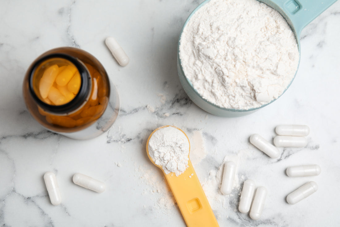 A bowl of white powder next to white supplement capsules and a dark brown supplement bottle, all on a marble countertop.