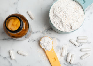 A bowl of white powder next to white supplement capsules and a dark brown supplement bottle, all on a marble countertop.