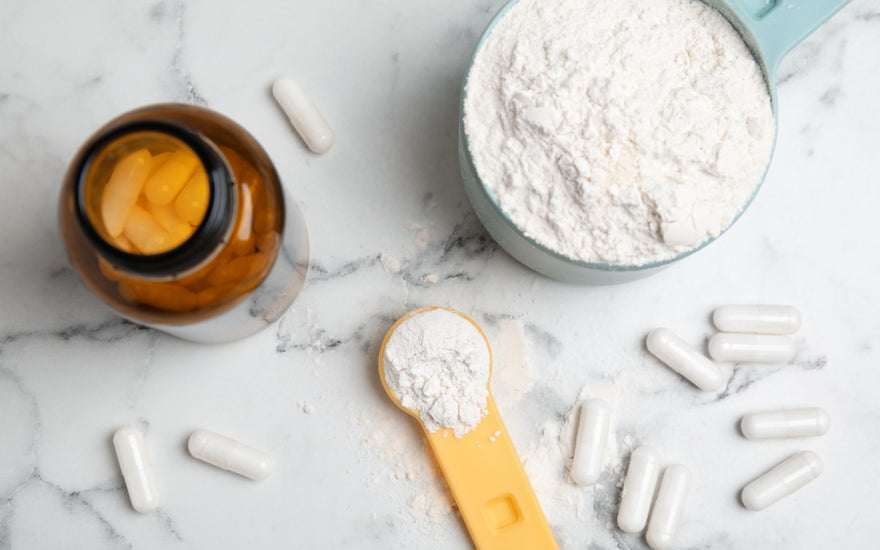 A bowl of white powder next to white supplement capsules and a dark brown supplement bottle, all on a marble countertop.