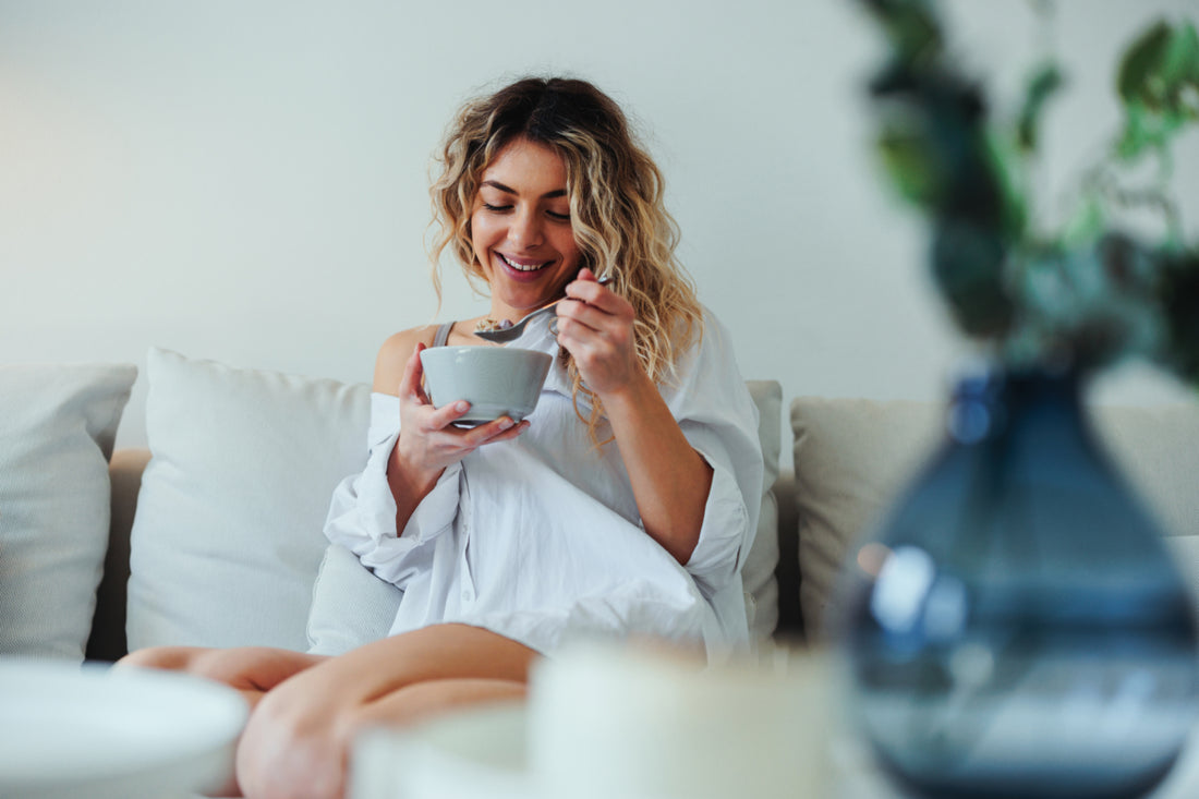 Woman in a white blouse sitting on a couch eating from a grey ceramic bowl.