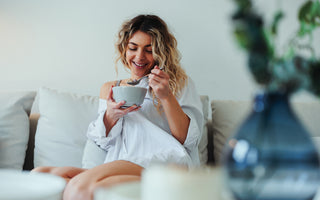 Woman in a white blouse sitting on a couch eating from a grey ceramic bowl.