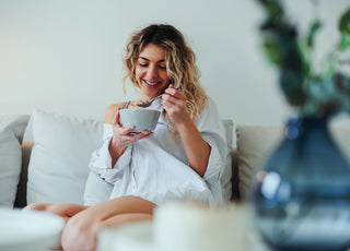 Woman in a white blouse sitting on a couch eating from a grey ceramic bowl.