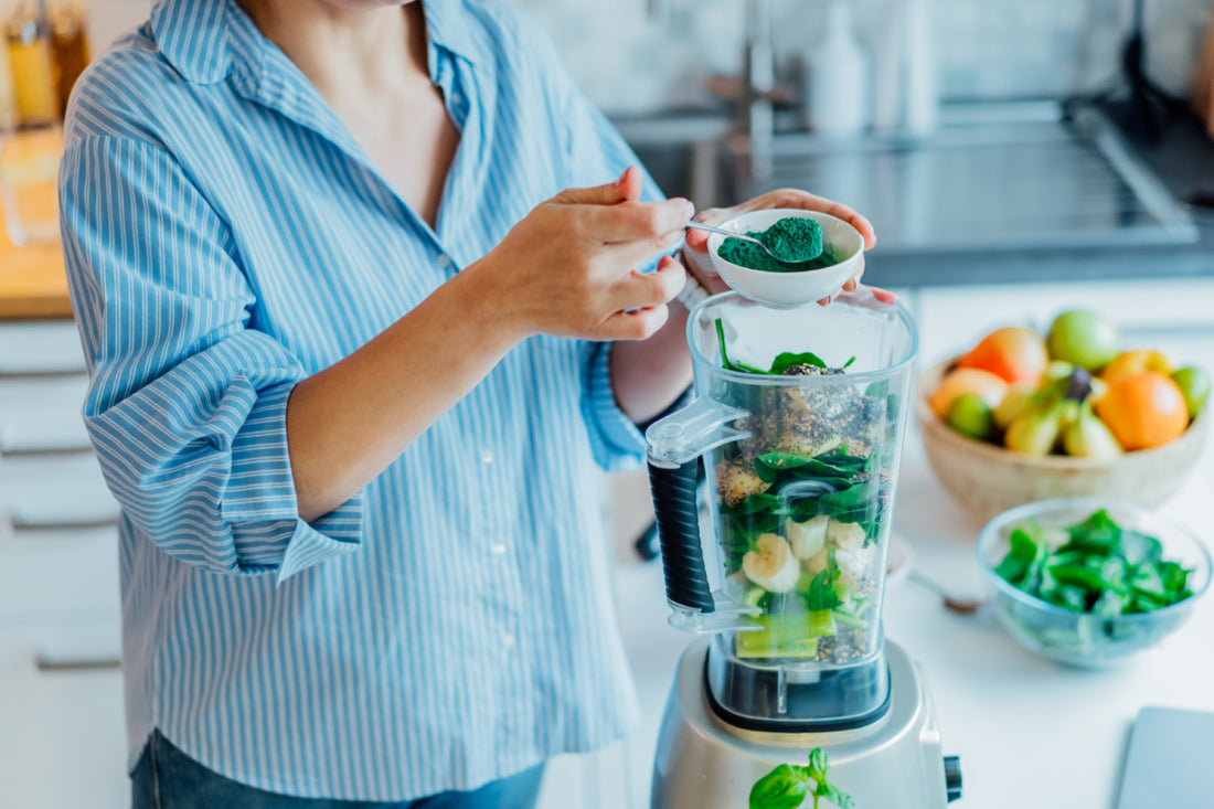 Woman in a blue striped shirt standing at the kitchen counter and scooping green powder into the blender that is already full of smoothie ingredients.