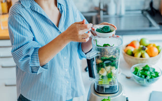 Woman in a blue striped shirt standing at the kitchen counter and scooping green powder into the blender that is already full of smoothie ingredients.