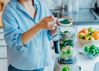 Woman in a blue striped shirt standing at the kitchen counter and scooping green powder into the blender that is already full of smoothie ingredients.