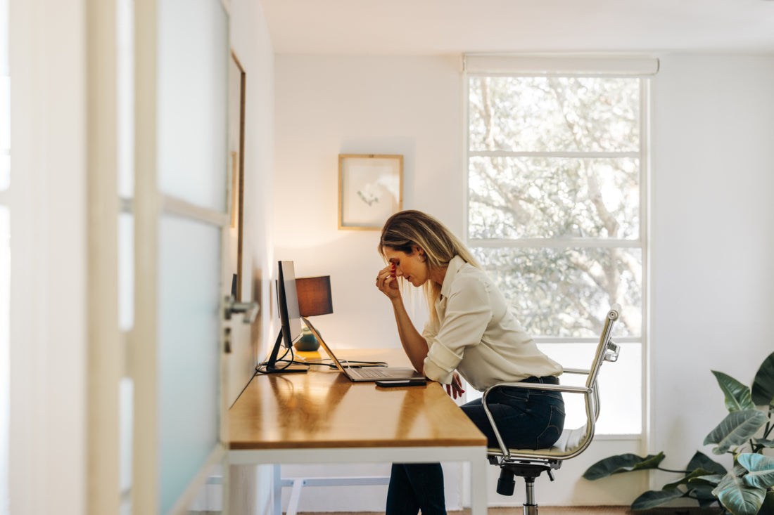 Woman in business casual attire sitting at a desk in front of her laptop and desktop. She has her fingers placed on the bridge of her nose and her head tilted down as if she is stressed.