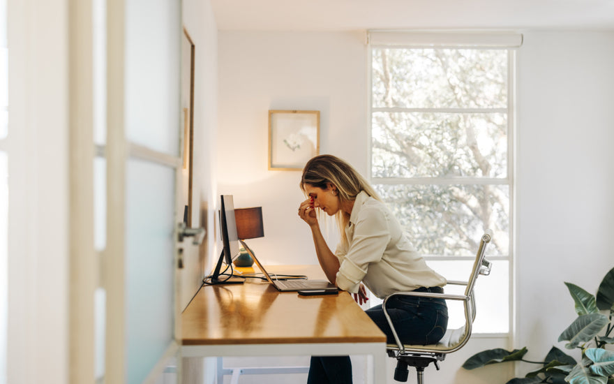 Woman in business casual attire sitting at a desk in front of her laptop and desktop. She has her fingers placed on the bridge of her nose and her head tilted down as if she is stressed.