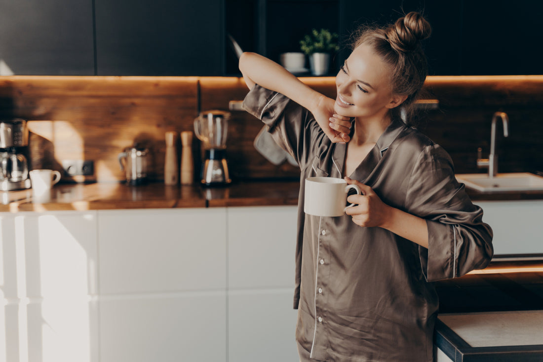 Relaxed woman in brown pajamas smiling and stretching in the kitchen while holding a cup of coffee.
