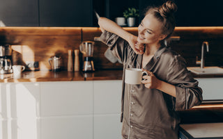 Relaxed woman in brown pajamas smiling and stretching in the kitchen while holding a cup of coffee.