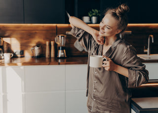 Relaxed woman in brown pajamas smiling and stretching in the kitchen while holding a cup of coffee.