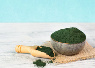 A bowl and scoop full of green algae powder sitting on a grey countertop in front of a light blue wall. 