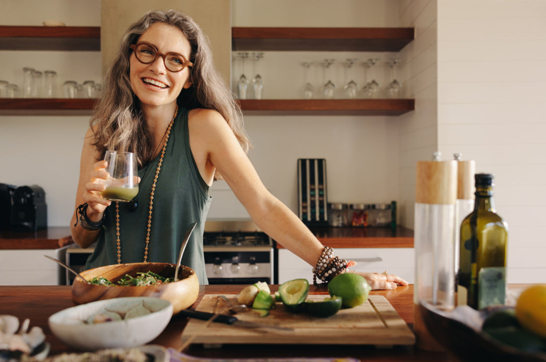 Middle-aged woman standing with one hand resting on the kitchen counter while she drinks a green smoothie out of a glass.