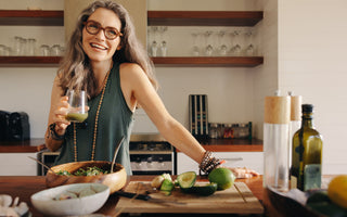 Middle-aged woman standing with one hand resting on the kitchen counter while she drinks a green smoothie out of a glass.