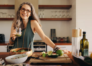 Middle-aged woman standing with one hand resting on the kitchen counter while she drinks a green smoothie out of a glass.