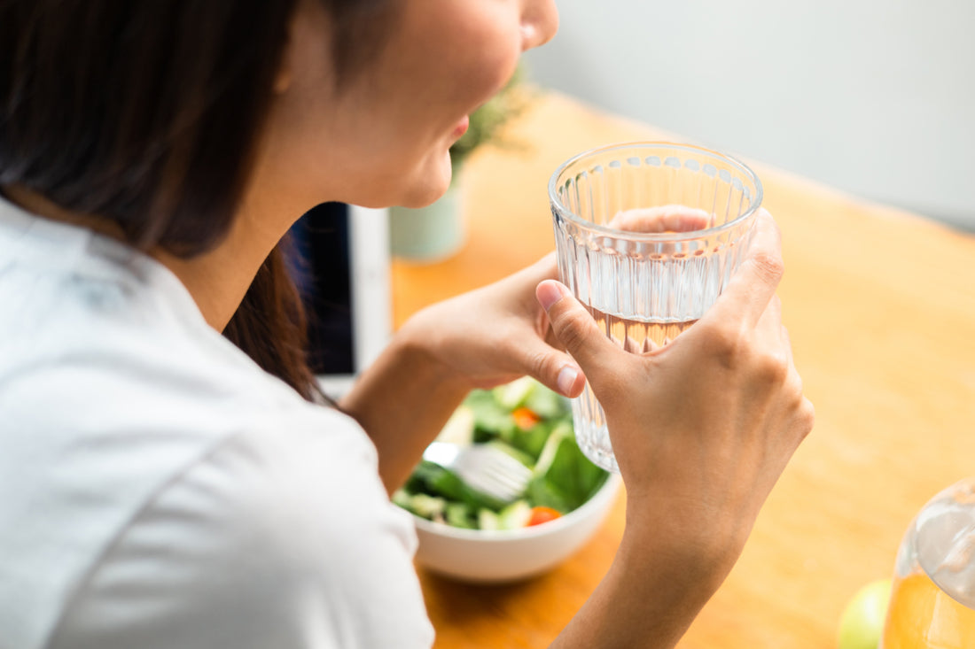 Woman sitting at a table with her lunch. She is holding a glass of water between both hands.