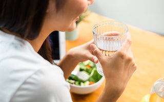 Woman sitting at a table with her lunch. She is holding a glass of water between both hands.