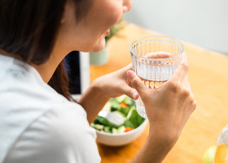 Woman sitting at a table with her lunch. She is holding a glass of water between both hands.