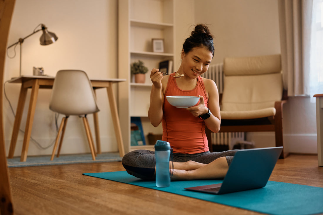 Woman in workout clothes sitting cross-legged on a blue yoga mat in her living room eating a bowl of yogurt while looking at something on her laptop, which is open on the mat in front of her. 