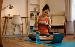 Woman in workout clothes sitting cross-legged on a blue yoga mat in her living room eating a bowl of yogurt while looking at something on her laptop, which is open on the mat in front of her. 