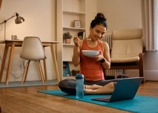 Woman in workout clothes sitting cross-legged on a blue yoga mat in her living room eating a bowl of yogurt while looking at something on her laptop, which is open on the mat in front of her. 