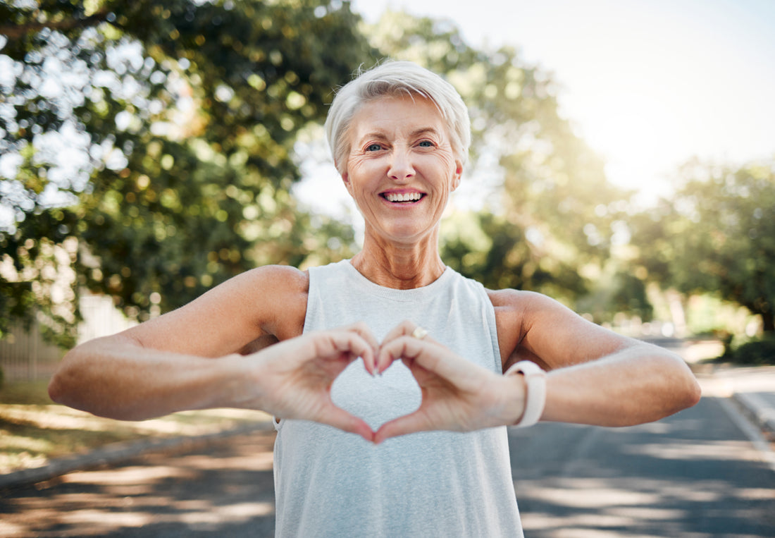 Older woman with short white hair is wearing a white tank top while standing on a road with her hands held out in front of her to make a heart.