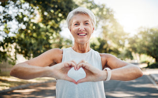 Older woman with short white hair is wearing a white tank top while standing on a road with her hands held out in front of her to make a heart.