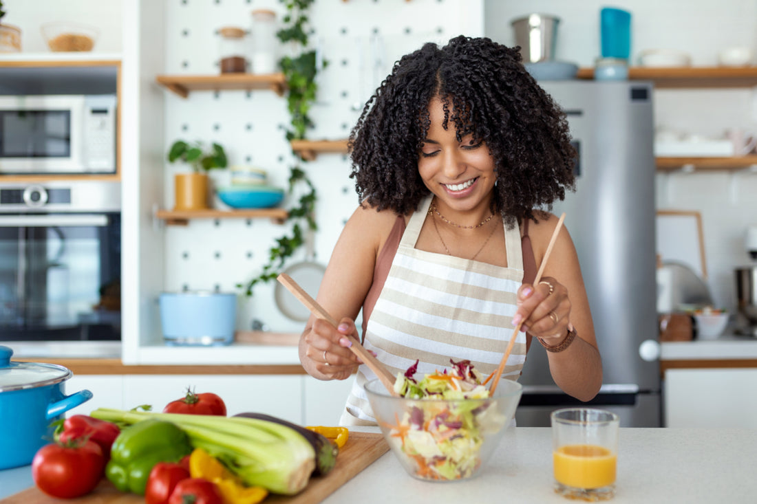 Woman wearing a striped apron using wooden utensils to mix a salad in a glass bowl at the kitchen counter.