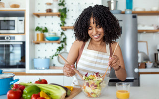 Woman wearing a striped apron using wooden utensils to mix a salad in a glass bowl at the kitchen counter.