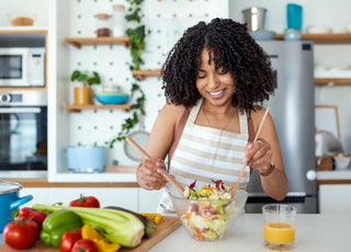 Woman wearing a striped apron using wooden utensils to mix a salad in a glass bowl at the kitchen counter.