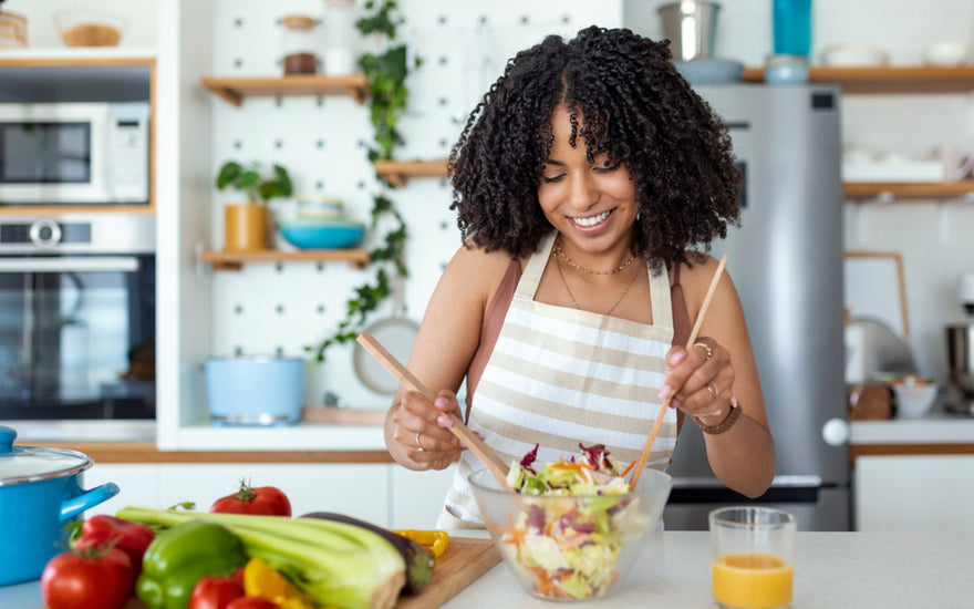 Woman wearing a striped apron using wooden utensils to mix a salad in a glass bowl at the kitchen counter.