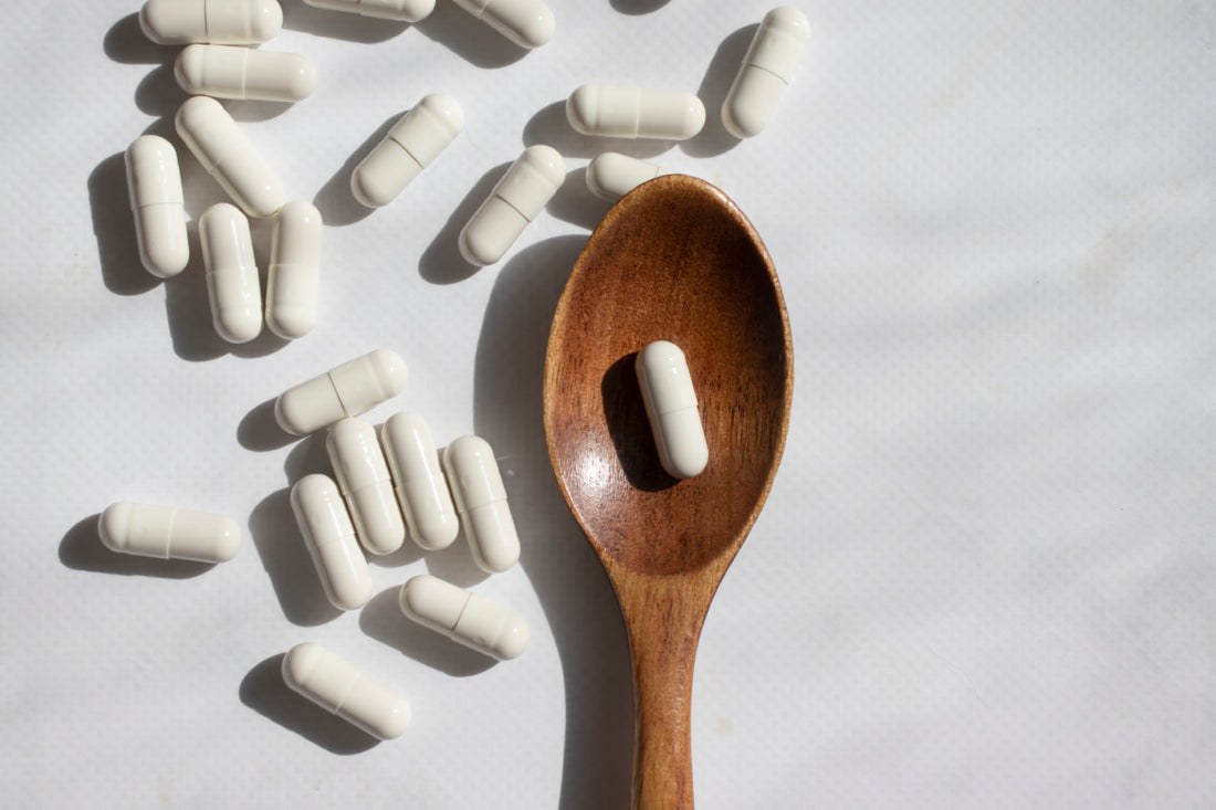 White supplement capsules scattered around a wooden spoon that contains a single capsule.