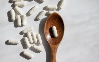 White supplement capsules scattered around a wooden spoon that contains a single capsule.