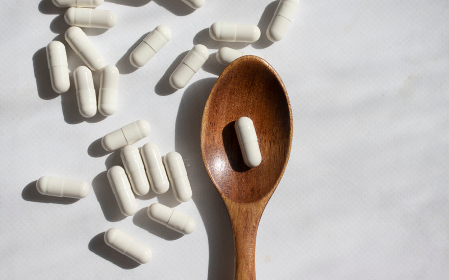 White supplement capsules scattered around a wooden spoon that contains a single capsule.