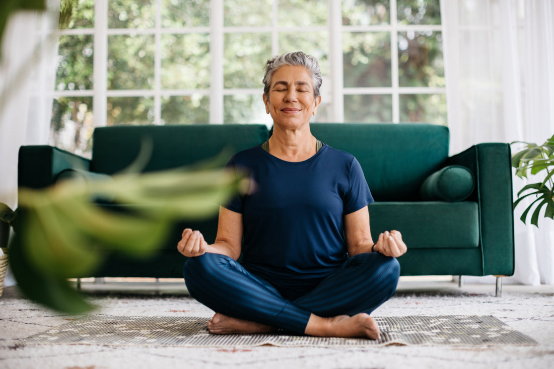 Middle-aged woman in all blue sitting cross-legged in a meditation position in front of a green couch.
