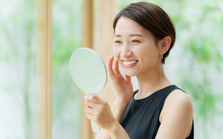 Adult female smiling while examining her skin in a handheld mirror.