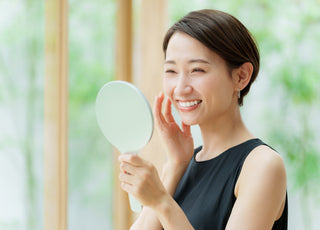 Adult female smiling while examining her skin in a handheld mirror.