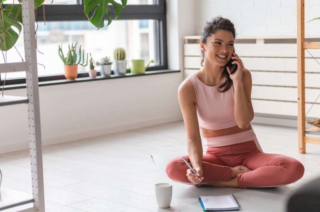 Happy young adult woman wearing pink fitness clothes is sitting on the floor talking on her cell phone and writing in a notebook. 