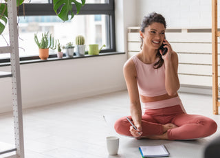 Happy young adult woman wearing pink fitness clothes is sitting on the floor talking on her cell phone and writing in a notebook. 