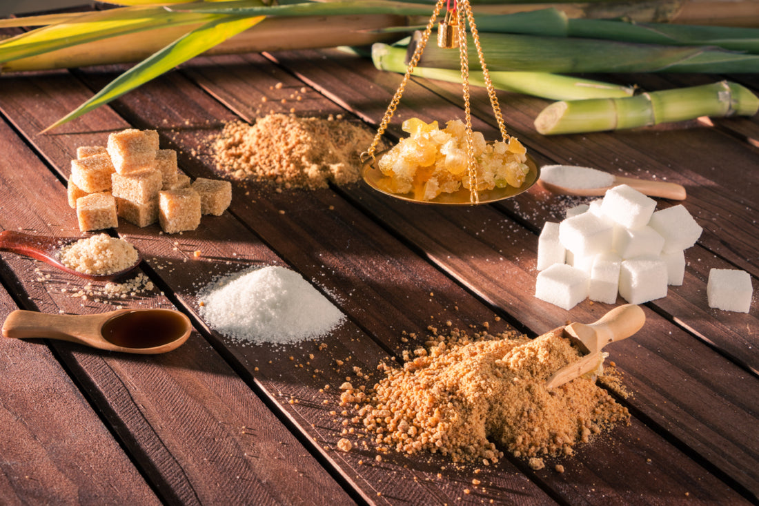 An array of different types of sugars, including brown sugar, honey clumps, and sugar cubes, sitting on a wooden table.