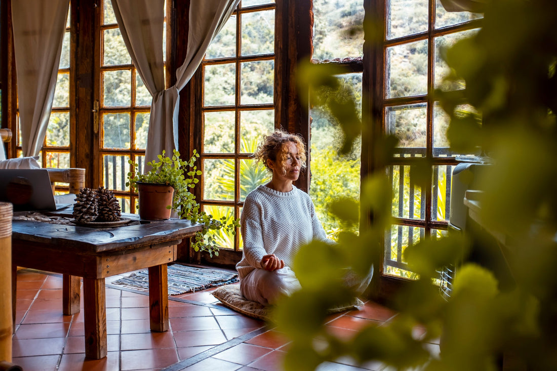 Woman sitting cross-legged on the floor meditating in front of a wall of large windows.