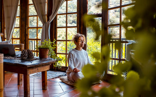 Woman sitting cross-legged on the floor meditating in front of a wall of large windows.