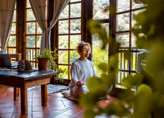 Woman sitting cross-legged on the floor meditating in front of a wall of large windows.