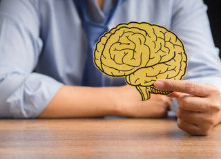 Person sitting at a table holding a yellow piece of paper with a brain drawn on it.