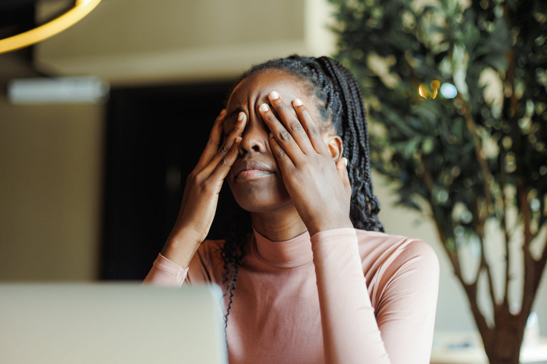Tired young woman covering her eyes with her hands while sitting in front of her laptop.
