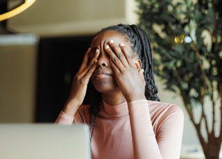 Tired young woman covering her eyes with her hands while sitting in front of her laptop.