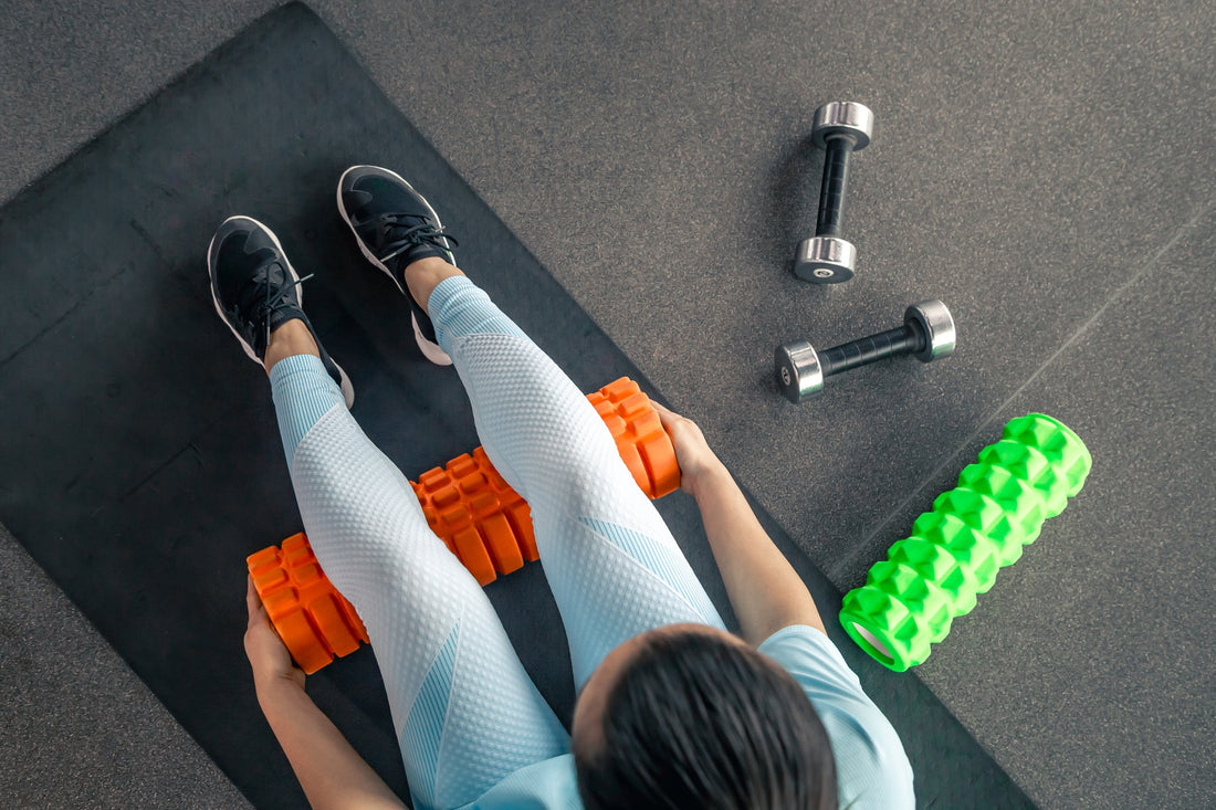 Woman wearing workout clothing sitting on a yoga mat using a foam roller on her legs.