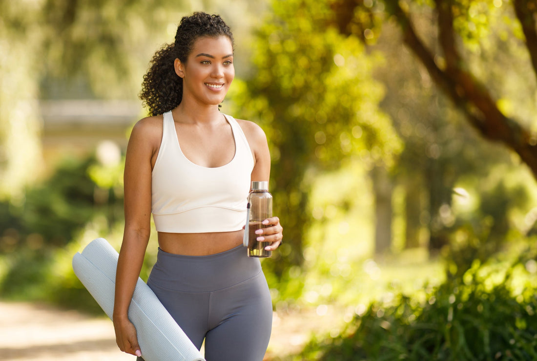 Athletic young lady holding a glass water bottle and gym mat while walking outside. 