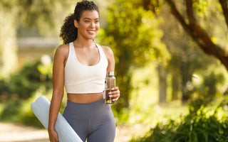 Athletic young lady holding a glass water bottle and gym mat while walking outside. 