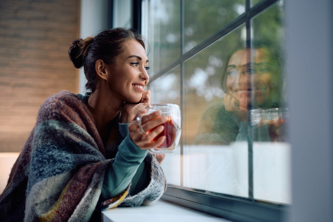 Young smiling woman drinking tea and looking through a window at home.